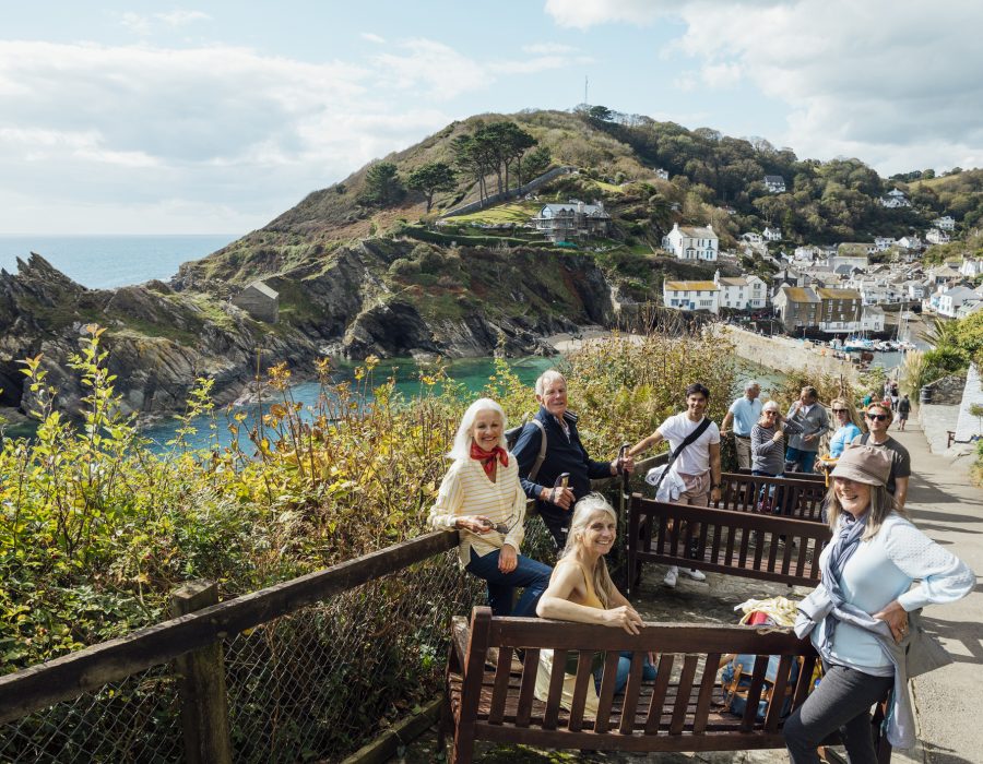 A group of male and female friends, of all ages, sitting on and standing around benches on a hill in Cornwall. They are looking at the camera and smiling with Polperro in the background.